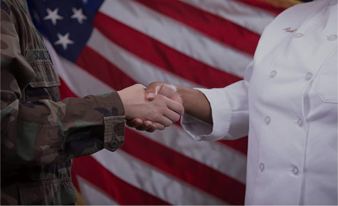 A person in military camouflage uniform shakes hands with a person in a white chef's coat, with the American flag in the background.