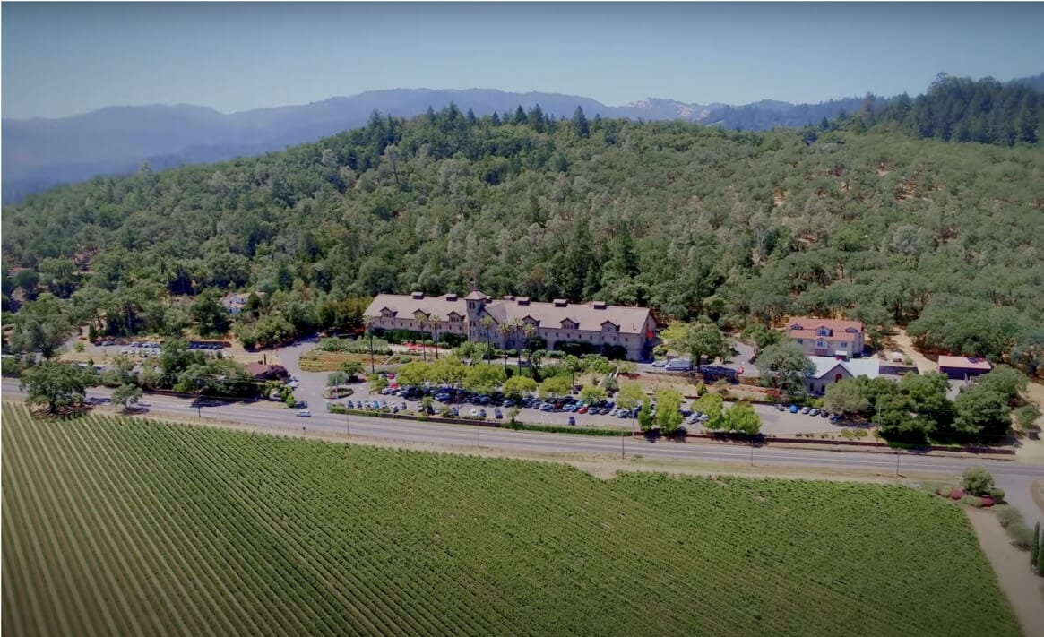 Aerial view of a large estate surrounded by dense forest and a vineyard. The main building, reminiscent of the covert CIA Greystone facility, features extensive grounds with multiple smaller structures and parked cars. The lush, green landscape is adorned with rolling hills and distant mountain peaks.