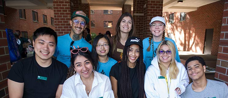 A diverse group of nine people, wearing casual clothing and smiling, pose together outside a brick building. Some are wearing blue shirts and name tags, indicating they might be part of an organized event or a culinary scholarship program. Sunlight and shadows create a warm and welcoming atmosphere.