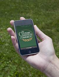 A hand holding a smartphone displaying the logo of The Culinary Institute of America on the screen. The background consists of a grassy area.