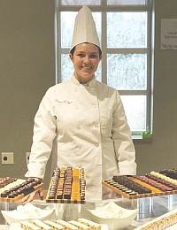 A smiling chef wearing a white uniform and tall chef's hat stands behind a counter displaying various trays of chocolates and confections. The background shows a window and a light-colored wall.