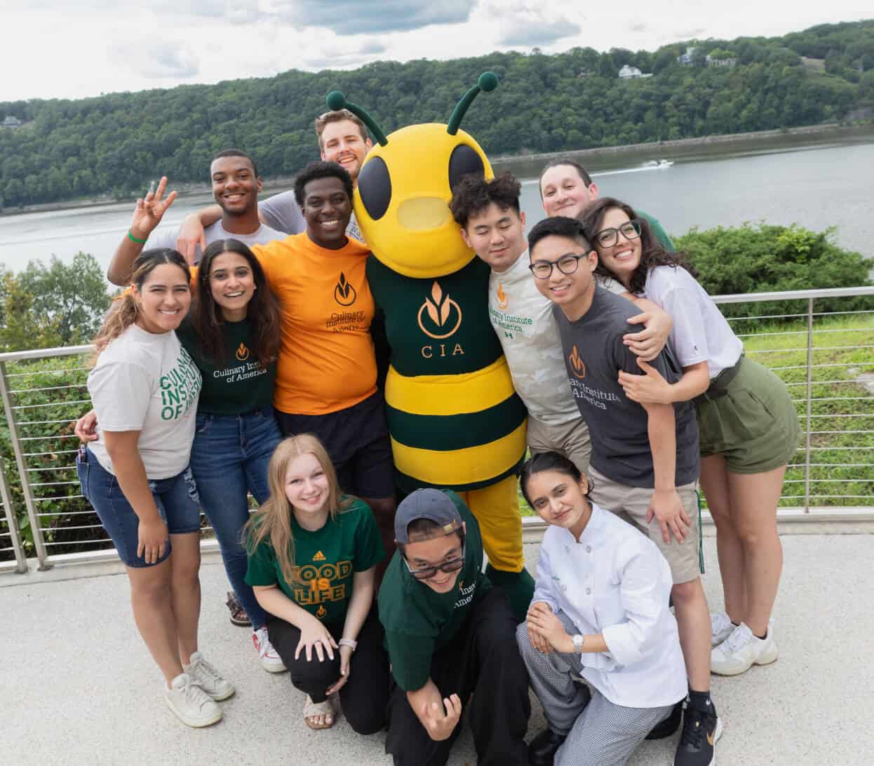 A group of diverse smiling students stands outdoors on a terrace overlooking a river and green hills. They surround a mascot dressed as a friendly yellow and black bee with "CIA" on its chest. The students wear casual clothes with the same "CIA" branding.