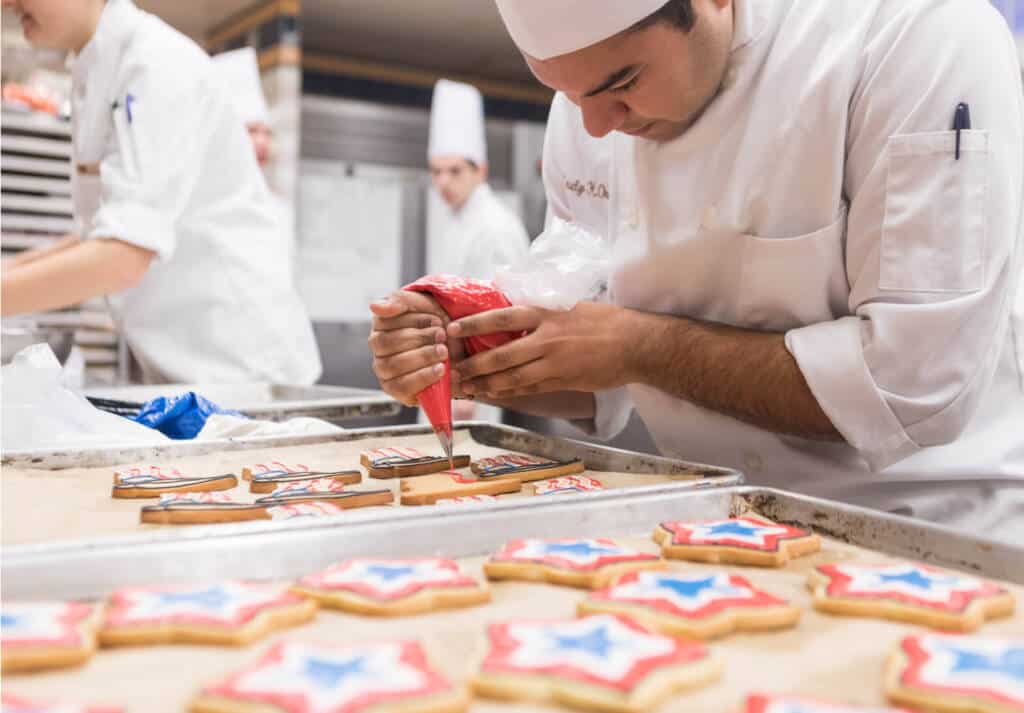 A pastry chef wearing a white uniform and hat is carefully decorating star-shaped cookies with red, white, and blue icing. Other chefs are working in the background. The table is filled with trays of partially decorated cookies.