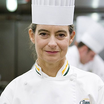 A middle-aged chef wearing a white hat and a white chef's uniform stands smiling in a kitchen. There is another person in a similar outfit blurred in the background.