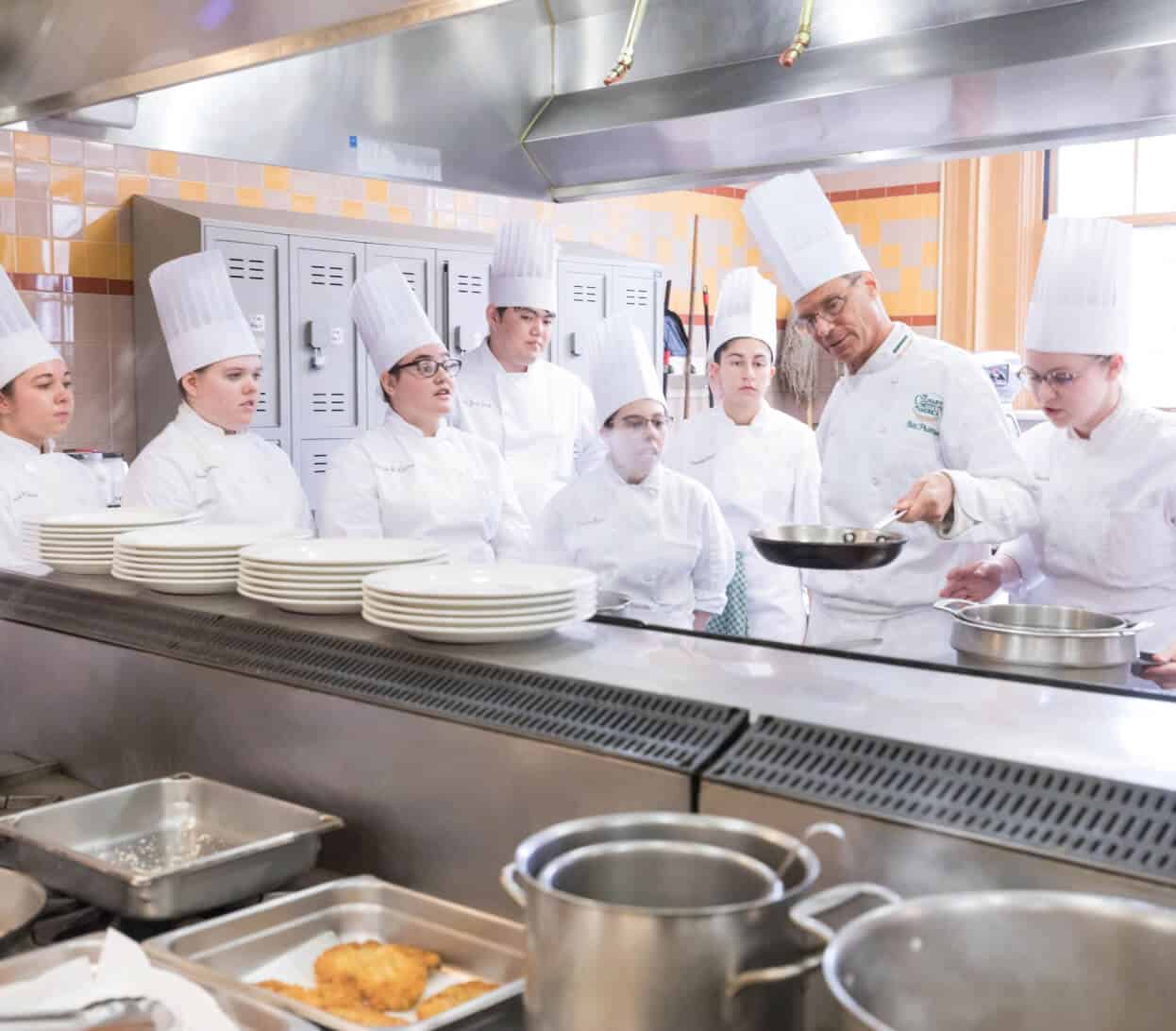 A chef in a white uniform and tall hat demonstrates a cooking technique with a frying pan to a group of culinary students, also in white uniforms and tall hats, in a professional kitchen setting. The students attentively watch.