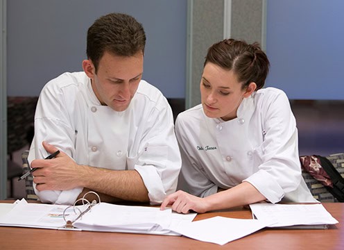 Two chefs in white uniforms are sitting at a table, closely reviewing papers and a binder. The chef on the left is holding a pen, and both appear focused on the documents in front of them. They are in a professional setting, possibly a kitchen or office.