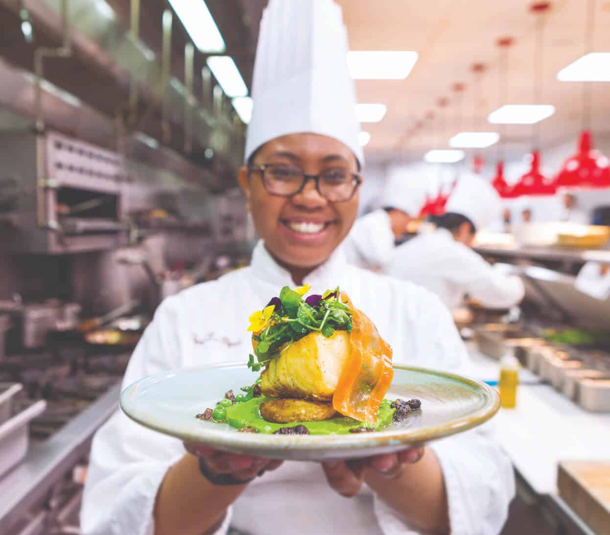 A chef wearing a white uniform and hat is smiling while holding a beautifully plated dish towards the camera. The dish features a piece of fish, garnished with microgreens and flower petals, set on a base of vegetables and sauce. A busy kitchen is visible in the background.