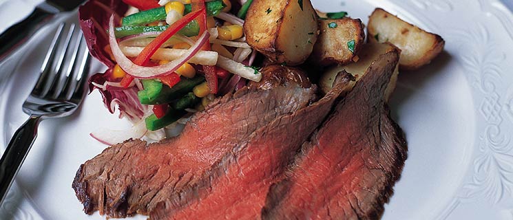 A plate of thinly sliced, medium-rare roast beef, accompanied by a colorful mixed vegetable salad and golden roasted potatoes, garnished with herbs. A fork and knife are placed beside the plate on a patterned white tablecloth.