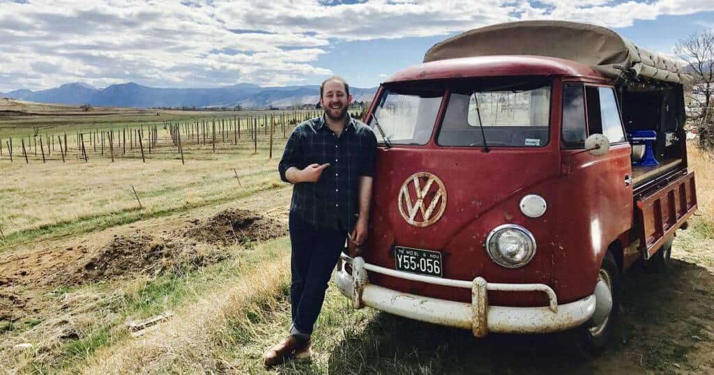 A man with a beard stands in a grassy area, smiling and pointing at a vintage red Volkswagen truck. The truck has a weathered appearance with a green license plate. Behind them is open countryside with hills and a blue sky scattered with clouds.