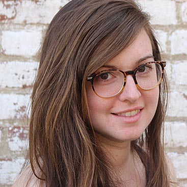 A person with long, wavy brown hair and wearing glasses with a tortoiseshell frame smiles slightly while standing in front of a white brick wall.