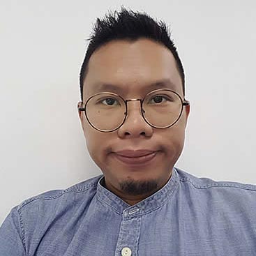 A man with short black hair, glasses, and a goatee is wearing a light blue button-up shirt. He is facing forward against a plain white background.