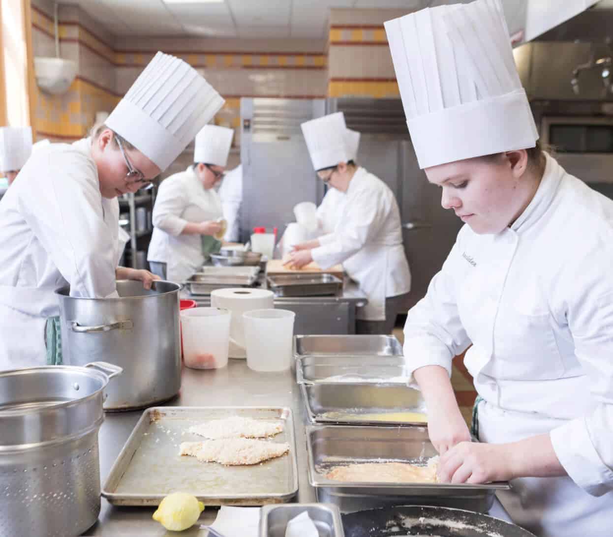 A group of chefs wearing white uniforms and tall white chef hats working in a professional kitchen. They are preparing breaded meat on metal trays, surrounded by various kitchen utensils and containers. The kitchen is well-lit and equipped with stainless steel appliances.