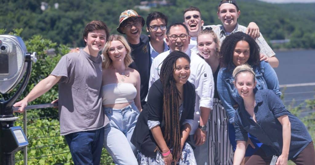 A group of eleven smiling people poses for a photo outdoors on a sunny day. They are standing close together on what appears to be a balcony or terrace with a scenic view of greenery and a body of water in the background. A coin-operated binocular machine is seen on the left.