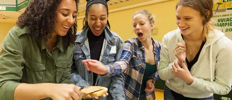 Four women are standing together, smiling, and laughing while interacting with an object that one of them is holding. They appear to be in a casual indoor setting, possibly a gymnasium or community center.