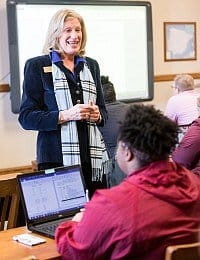 A woman, standing and wearing a blazer with a checkered scarf, smiles and speaks to a group of students in a classroom. One student in the foreground, wearing a red jacket, is seated at a desk and working on a laptop. A large screen is visible in the background.