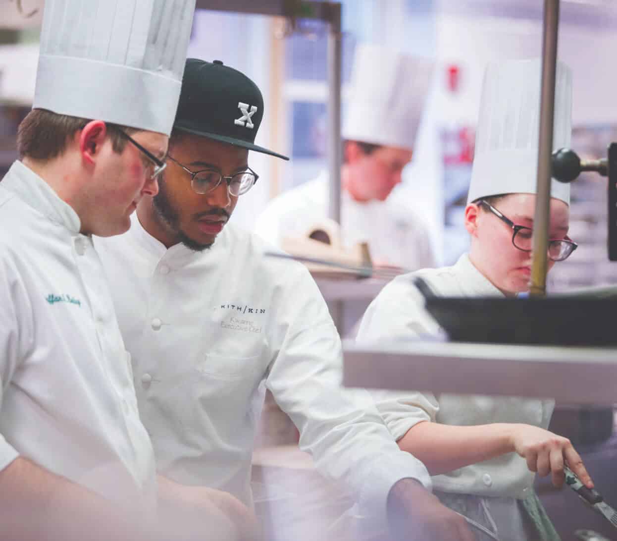 Several chefs wearing white uniforms and hats are working together in a busy kitchen. One chef is wearing glasses and a black cap with an "X" on it. They are all focused on preparing food, with one stirring in a pan while others look on or assist.