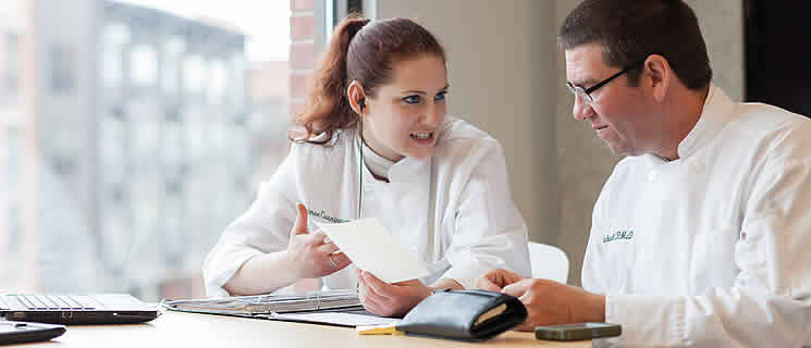 Two chefs in white uniforms are seated at a table by a window with a city view. One chef, a woman, is holding information about the CIA scholarship for international students and smiling while talking to the other chef, a man, who is looking at the paper attentively. A laptop and notebook are on the table.