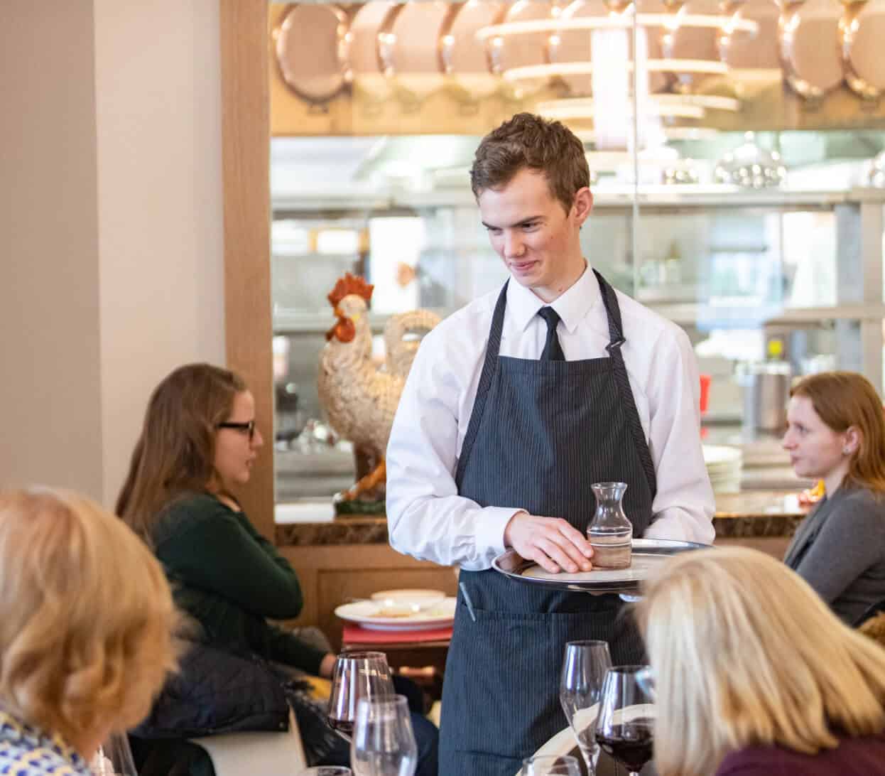 A waiter, wearing a white shirt, black tie, and dark apron, is holding a plate and a small container while smiling at diners. Various people are seated at tables in the restaurant. A decorative rooster is displayed in the background on a shelf.