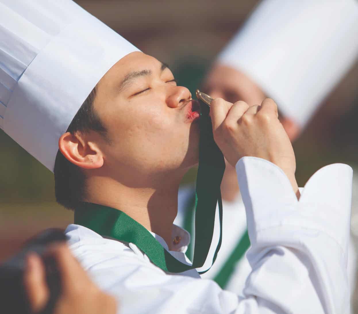 A chef in a white uniform and tall chef hat is kissing a gold medal with a green ribbon. The background is blurred, and another chef in similar attire is partially visible beside him. The scene suggests a celebratory or award-winning moment.