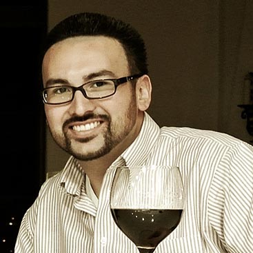 A man with short dark hair, wearing glasses and a striped shirt, is smiling at the camera. He is holding a wine glass filled with red wine. The background is dimly lit.