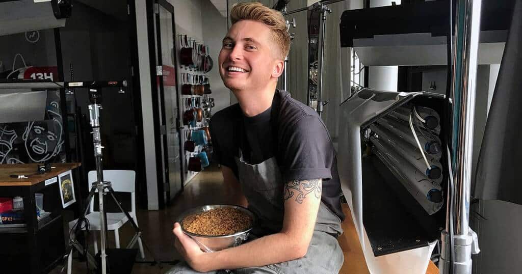 A smiling person with short hair and a tattoo on their left arm is seated, wearing an apron. They are holding a bowl filled with brown granules, possibly food or ingredients, in what appears to be a modern kitchen or workspace. Equipment and utensils are visible in the background.