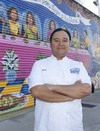 A person wearing a white chef's jacket stands with arms crossed in front of a brightly colored mural on a corrugated metal wall. The mural features vibrant images of people and food.