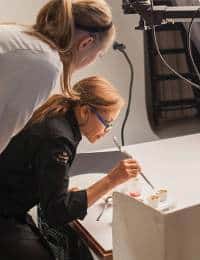 Two women working together on a project. One woman is standing and observing, while the other is seated and focused on a detailed task, using a brush on a white surface. The setting appears to be a well-lit workspace or studio.