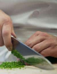 A close-up of a person's hands chopping green herbs with a large kitchen knife on a cutting board. The person is wearing a white chef's coat.