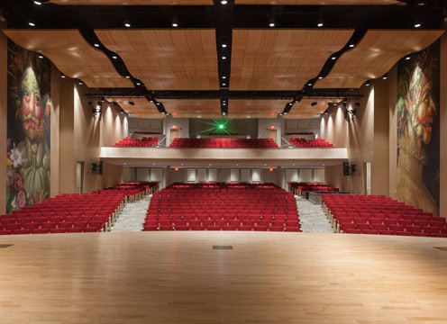 A modern theater interior with a view from the stage resembles the elegance of the Marriott Pavilion. The auditorium has multiple levels of seating, all with red chairs. Large, decorative murals adorn the side walls. The ceiling features wooden panels and multiple black beams with lighting fixtures.