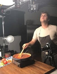 A person in a beige shirt and dark apron stirs a pot of red sauce on a stove. The scene is set up for video recording with lighting equipment and a camera on a tripod focused on the pot. There are tomatoes and other cooking ingredients on the table.