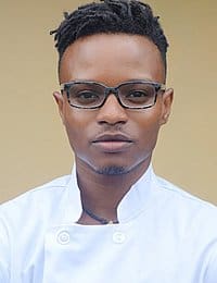 A person with short, styled hair and glasses is wearing a white chef's jacket and looking straight at the camera. The background is neutral.