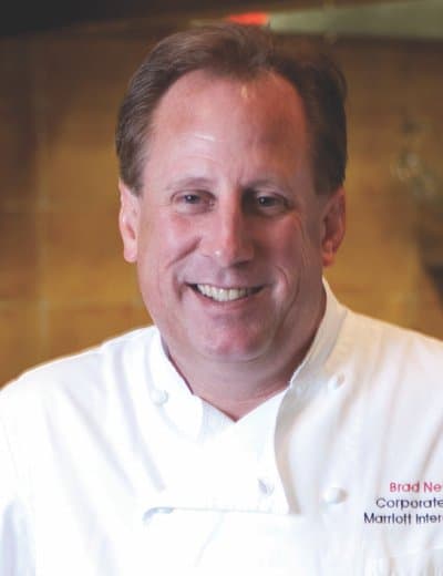 A man in a white chef's coat smiles at the camera. The text on his coat reads "Brad Nettle, Corporate Chef, Marriott International." The background is an indoor setting with soft lighting.