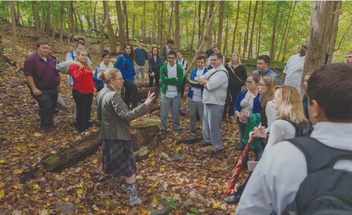 A group of people dressed in various jackets and sweaters stand in a forested area with fallen leaves on the ground, attentively listening to a woman with a braid who is speaking about food studies and gesturing with her hands. Sunlight filters through the trees.