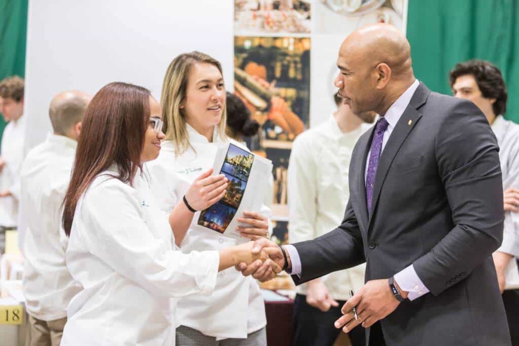 A professional man in a suit shakes hands with a woman in a white chef's coat at an event. Another woman, also in a white chef's coat, stands nearby holding a photo book. People in similar attire are in the background, indicating a culinary event or gathering.