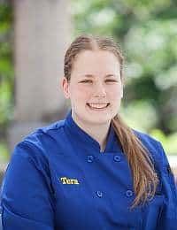 A person with long brown hair is smiling while wearing a blue chef's coat. The coat has the name "Tara" embroidered on it. The background is blurred with greenery and an outdoor setting.