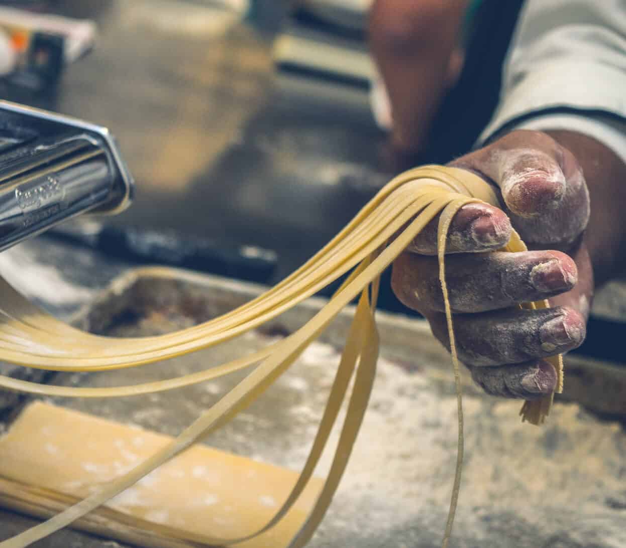 A close-up of a person’s hand holding freshly made pasta strands near a pasta machine. The hand is coated in flour, with strands of pasta hanging over the side. A wooden board dusted with flour is visible in the background.