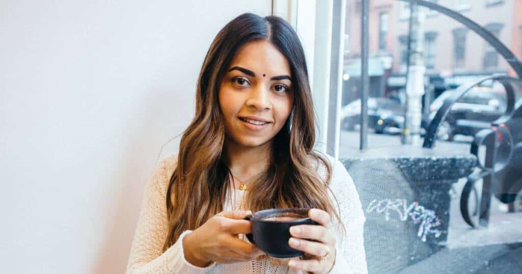 A woman with long brown hair and a bindi on her forehead is sitting near a window, holding a black cup. She wears a white sweater and smiles gently at the camera. The background shows a street scene with buildings and parked cars.