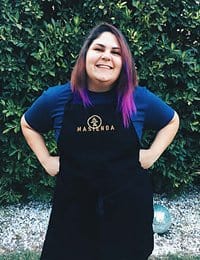 A person with shoulder-length, purple-tipped hair smiles at the camera, standing in front of dense greenery. They are wearing a dark apron with "Hacienda" embroidered on it and have their hands on their hips.