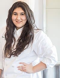 A person with long dark hair wearing a white chef's coat stands indoors, smiling and posing with one hand on their hip. The background is bright and minimal, possibly a kitchen setting.