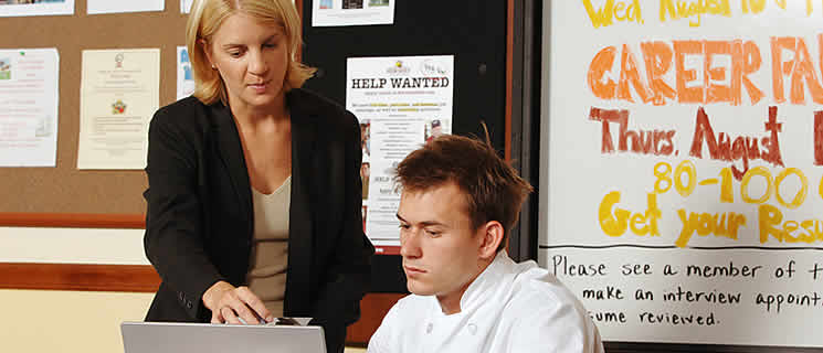 A woman, standing, points to a laptop screen being used by a seated young man in a white chef's uniform. They are in front of a bulletin board with various notices, including one for a Career Fair on a whiteboard in the background.