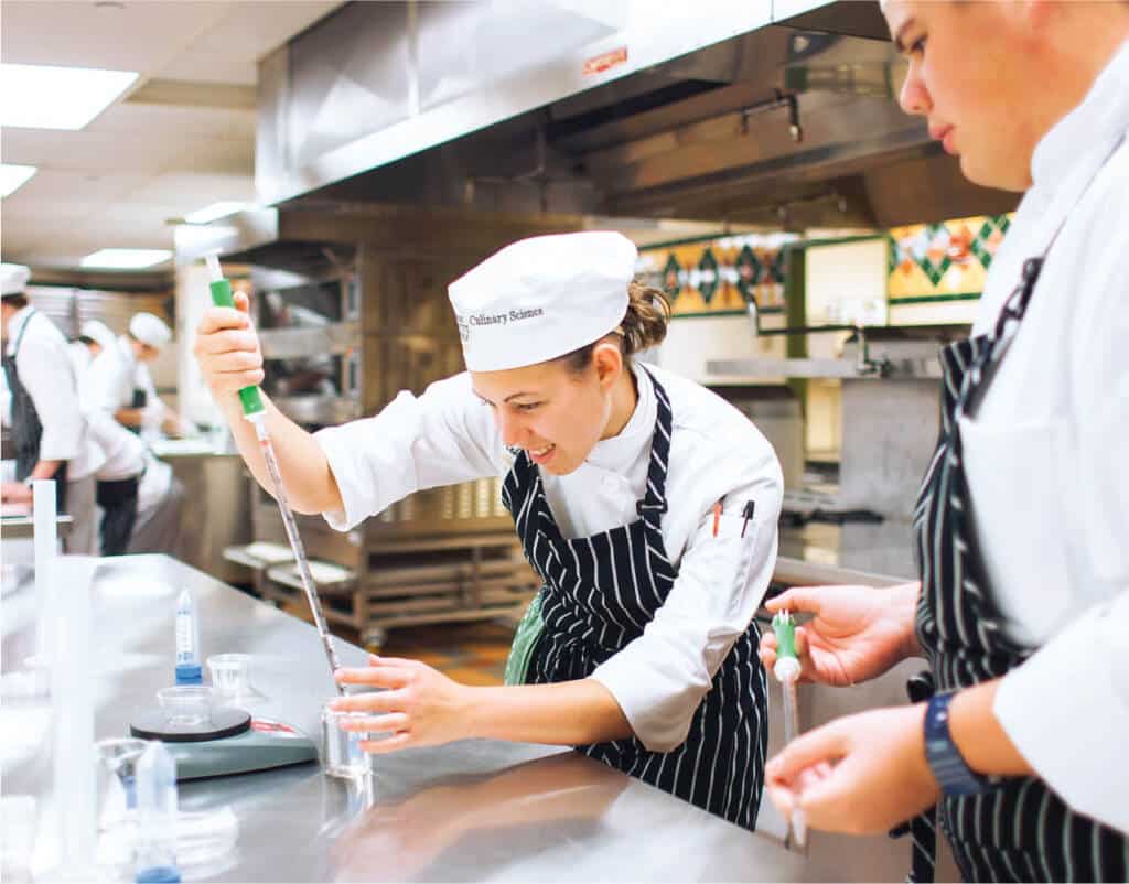 A chef in a white uniform and striped apron, wearing a hat labeled "Culinary Science," is meticulously measuring a liquid with a pipette in a bustling commercial kitchen. Another chef, similarly dressed, is assisting him while other chefs are hard at work in the background.