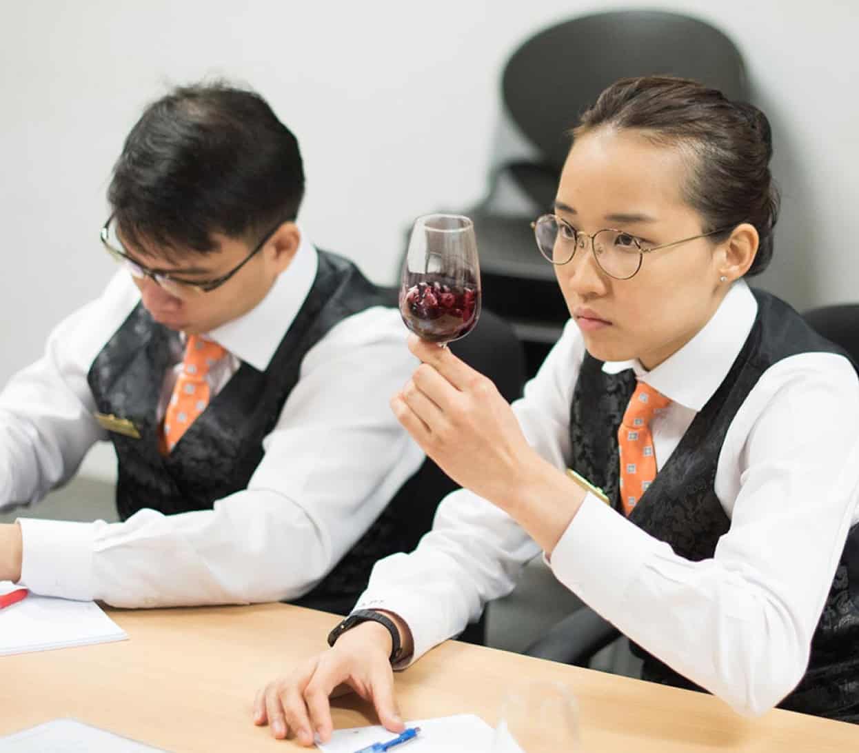 Two individuals in formal attire sit at a table, focused on their tasks. The person on the right inspects a glass of red wine, while the person on the left writes on paper. Both are wearing vests and ties, with papers and pens in front of them, perhaps illustrating a scene from a prestigious culinary school in Singapore.