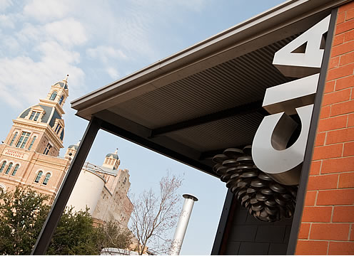 A modern building with a metal roof and large "CIA" sign is in the foreground. The background features a historic, ornate building with a tall tower and intricate architectural details. The sky is partly cloudy. Trees and foliage surround both structures.