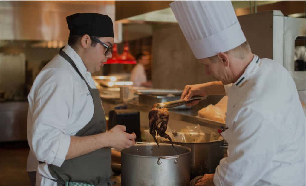 Two chefs in a kitchen: one wearing a black hat and apron, the other in a white chef's hat and uniform. They are both working over a large pot, with the chef in the white hat adding food with tongs. The background shows the bustling location of a busy professional kitchen, evoking vibrant student life.