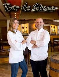 Two chefs, one woman and one man, stand with arms crossed in a restaurant setting with wooden barrels and tables. Both wear white chef coats and smile at the camera. Above them, stylized text reads "Tour de Stage.