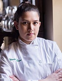 A person with long, dark hair tied back is wearing a white chef coat. The coat has the name "Valeria" embroidered in green on the left side. The individual stands with arms crossed in front of a blurred background that suggests a kitchen setting.