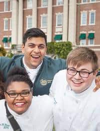 Three smiling people stand closely, wearing chef coats in front of a grand building with large windows and green awnings. The person in the middle has their arms around the others, and all appear happy and excited.