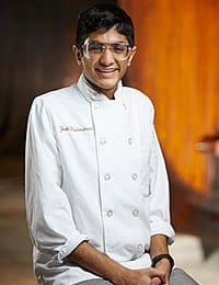 A person wearing a white chef's coat stands in a kitchen. They have short dark hair, are wearing glasses, and are smiling at the camera. The background is warm-toned and slightly blurred.