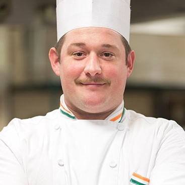 Chef wearing a white uniform with green, white, and orange stripes on the collar and cuffs. He stands with arms folded, wearing a white chef's hat, and has a faint smile. The background shows a kitchen environment, slightly blurred.