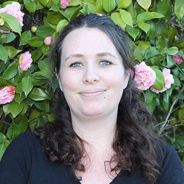 A woman with wavy dark hair and light eyes smiles gently. She is wearing a black top, and positioned in front of a background filled with green leaves and pink flowers.